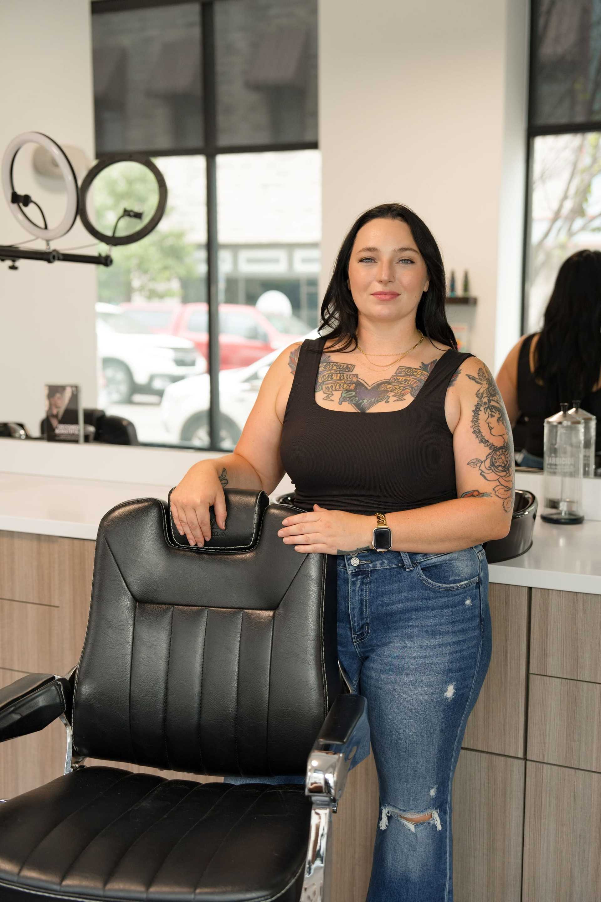 Female tattooed barber standing next to a salon chair in a modern barbershop.
